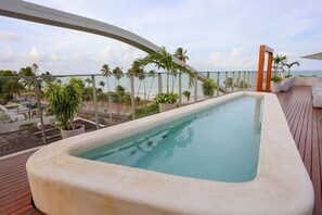 Una piscina en la azotea con vista al océano, rodeada de palmeras y sillones de descanso.