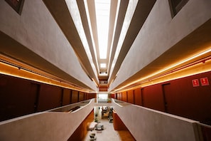 Un pasillo moderno de hotel con un pasillo central, barandillas de cristal e iluminación lineal en el techo.