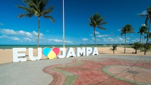Uma área pavimentada com um design colorido em formato de coração e palmeiras, que leva a uma praia com a inscrição "EU ♥ JAMPA".