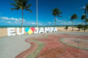 Un área pavimentada con un diseño colorido en forma de corazón y palmeras, que conduce a una playa con el texto 'EU ♥ JAMPA'.