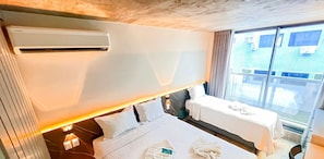 Una habitación de hotel con cama, mesita de noche, una pequeña mesa, unidad de aire acondicionado y ventana con vistas.