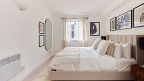 A bedroom with a bed, a radiator, a window with curtains, and framed artwork on the walls.