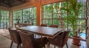 A spacious room with a large wooden table and chairs, large windows with a view of lush greenery, and a potted plant.