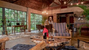 A spacious living area with a large wooden coffee table, a striped armchair, and a dining table with chairs. There is a large window providing a view of the outdoors. - Poolside 2-Bedroom Apartment with heated pool at the Jungle Oasis (Nairobi)