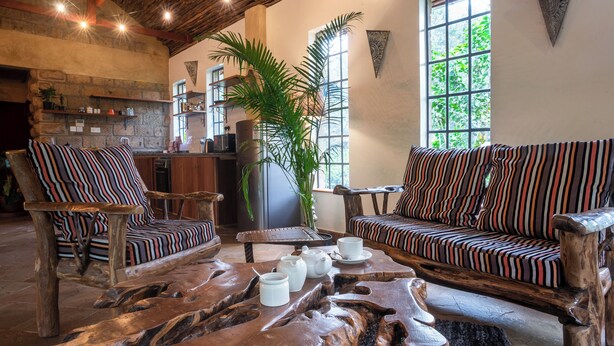 A cozy lounge area with striped sofas and chairs, a wooden coffee table, and a large window with a view of greenery. - Poolside 2-Bedroom Apartment with heated pool at the Jungle Oasis (Nairobi)