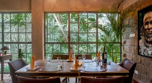 A dining area with a large window overlooking a lush garden, a wooden table set for a meal, and a wall-mounted painting.