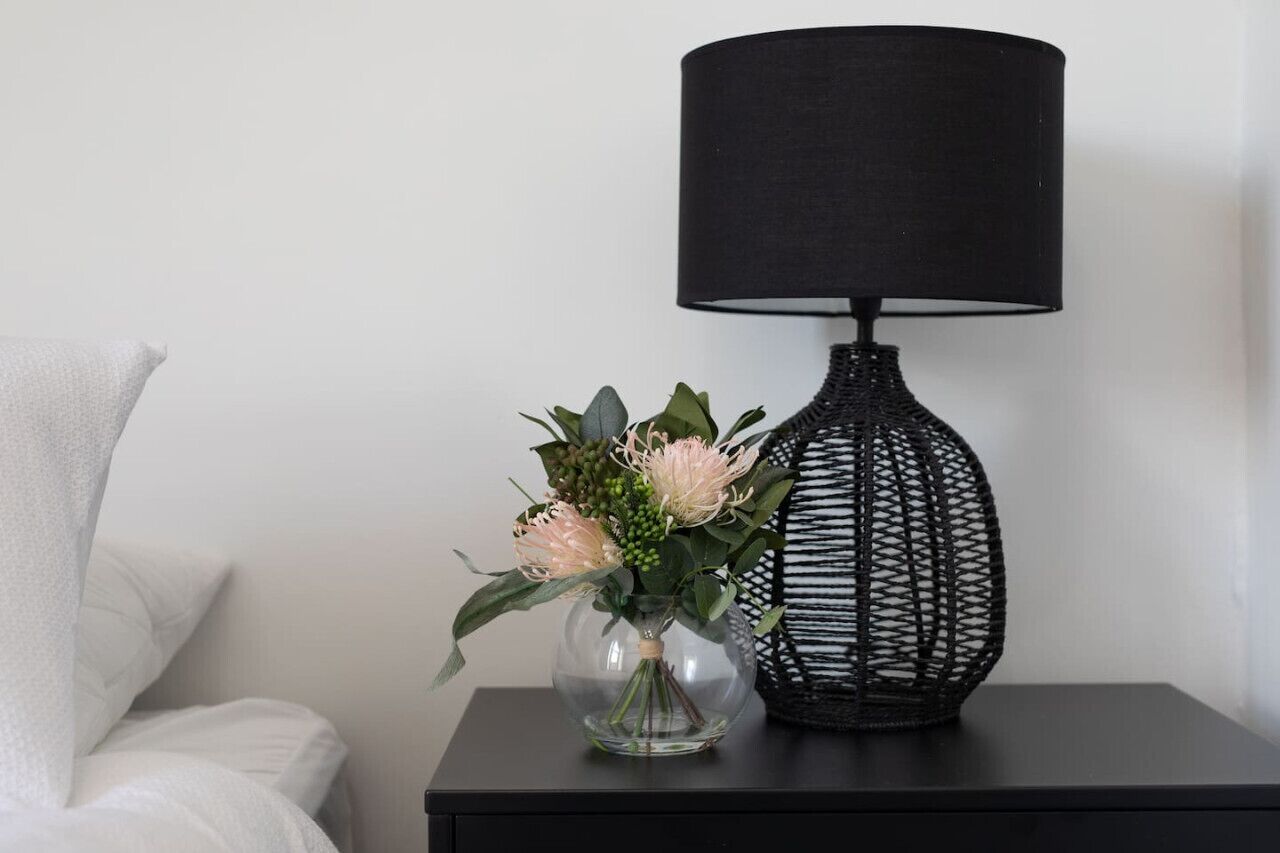A black lamp with a woven base on a black nightstand next to a clear vase with flowers.