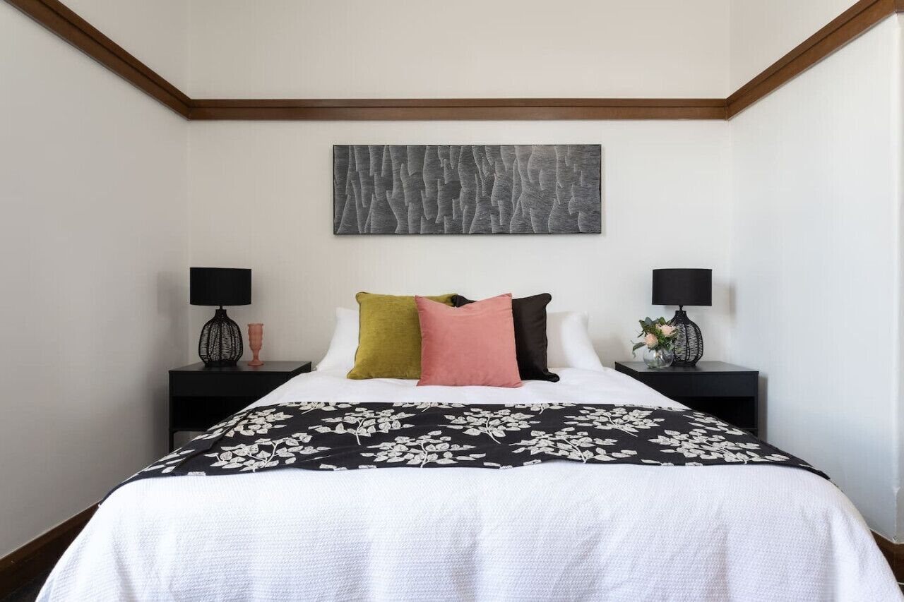 A bedroom with a bed, two bedside tables, a black and white floral bedspread, and a gray abstract painting on the wall.