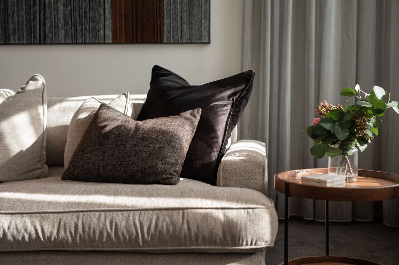 A modern living room with a beige sofa, a small round wooden side table, and a vase with green foliage.