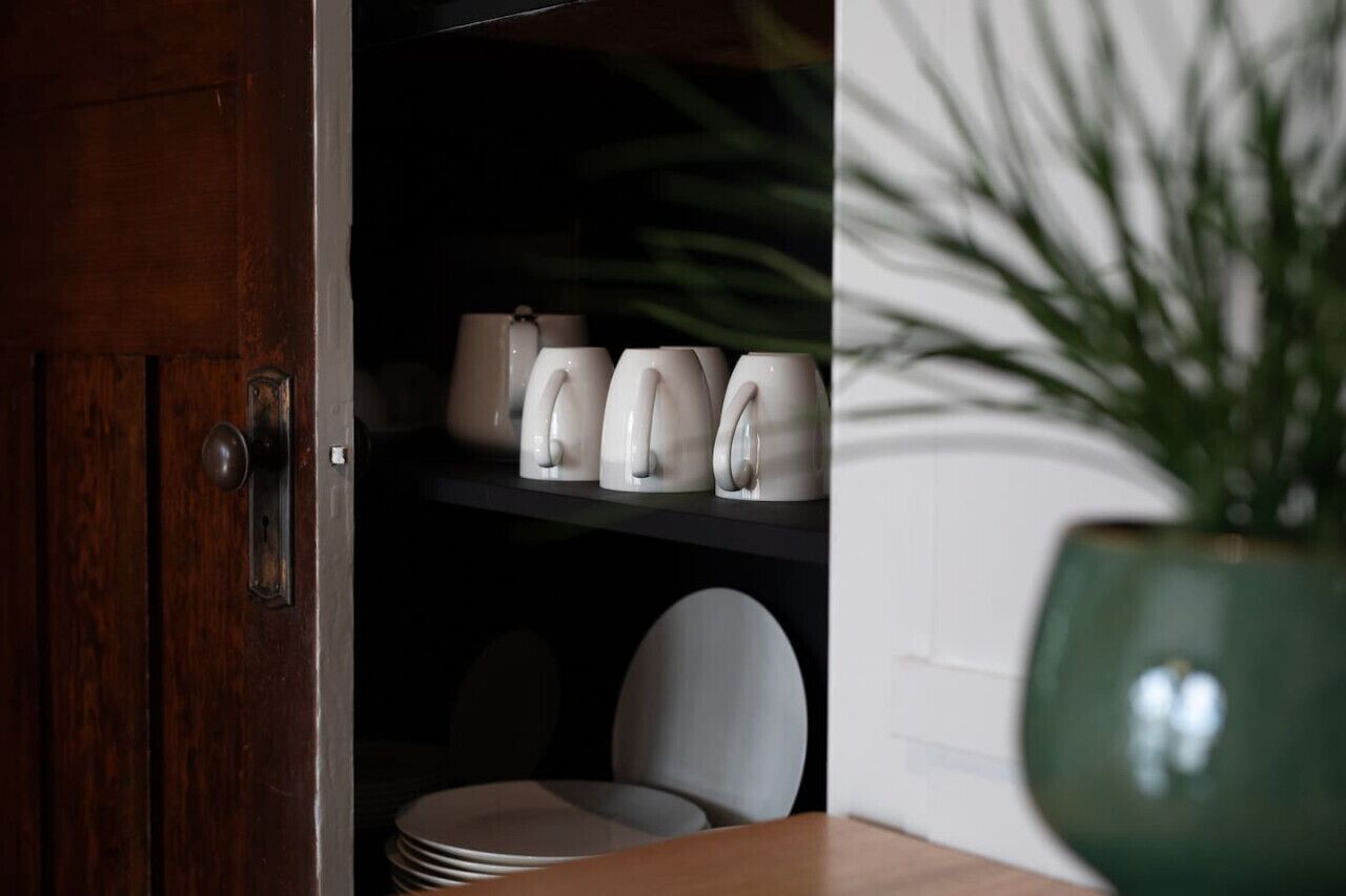 Een keukenhoek met een houten deur, een plank met witte mokken en een groene plant in pot.