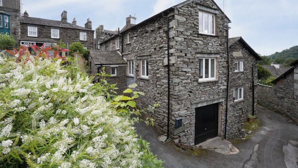 A stone building with a black door and multiple windows, surrounded by greenery and other stone buildings.
