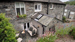 A stone house with a sloped roof, a small patio with outdoor furniture, and a garden with flowering plants.