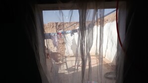 Vista a través de una ventana con una cortina ligera, mostrando un tendedero con ropa, una silla y un neumático.