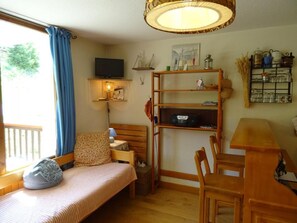 Quarto aconchegante com uma cama, uma escrivaninha e uma cadeira. Há uma estante com diversos objetos e uma janela com cortinas.