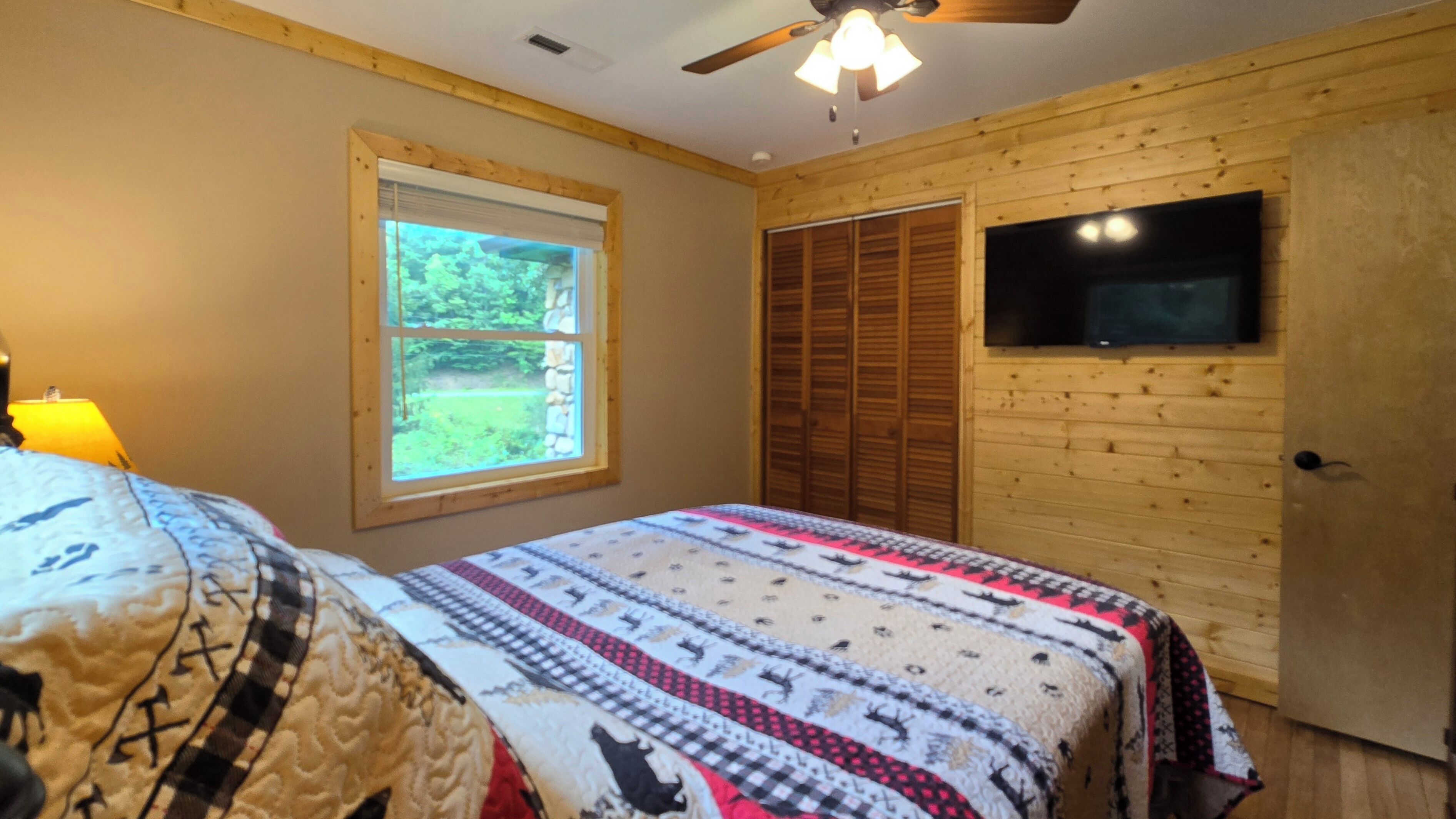 A bedroom with a bed, a window, a ceiling fan, and a TV mounted on the wall.
