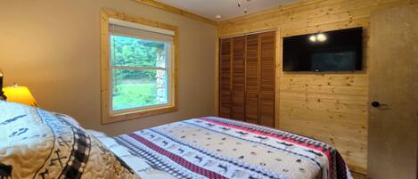 A bedroom with a bed, a window, a ceiling fan, and a TV mounted on the wall.