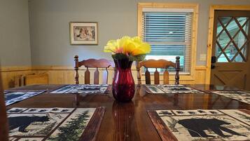 A dining table with a red vase and yellow flowers, surrounded by wooden chairs and patterned placemats.