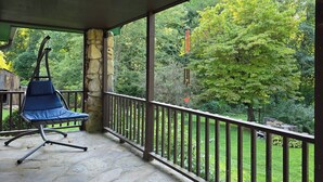 A covered outdoor area with a stone pillar, a blue chair, and a metal railing overlooking a garden with trees and a wooden structure. - Yellow Creek Cabin (Robbinsville)