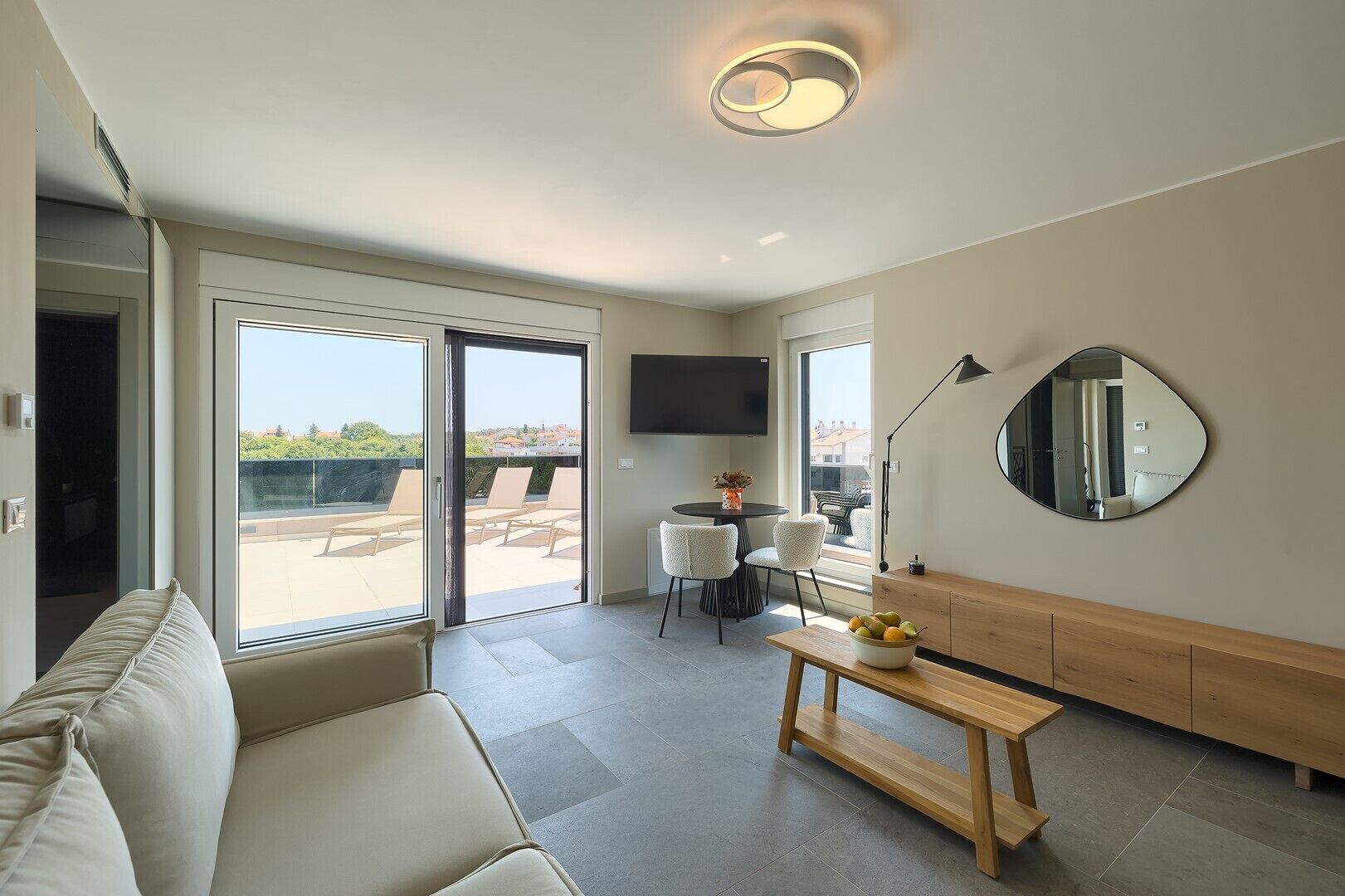 A modern living room with a sofa, a dining table with chairs, a TV mounted on the wall, and a balcony with outdoor seating.