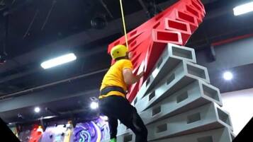 Une personne escalade un mur d’escalade aux motifs géométriques, de couleurs rouge et grise.