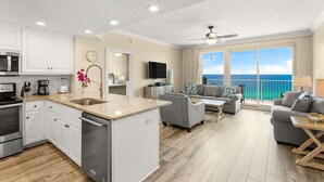 A modern kitchen with white cabinets, stainless steel appliances, and a large island. There is a living area with a gray sofa and a view of the ocean through large windows. - Gulf-front escape w/ tickets, pool & hot tub (Panama City Beach)