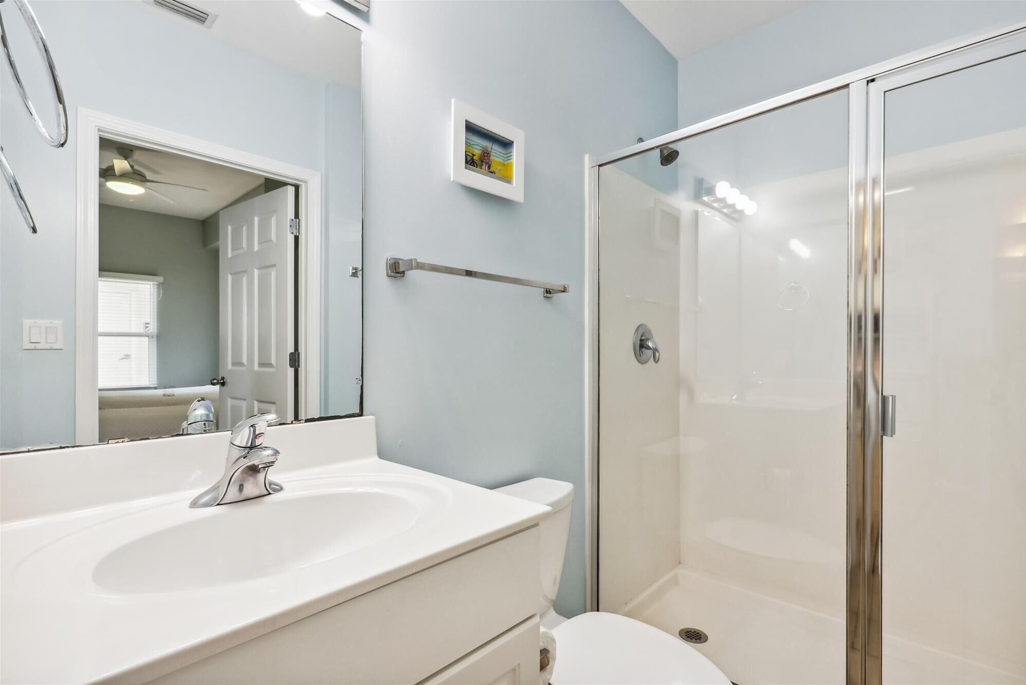 Een badkamer met een wit wastafelmeubel, een spiegel, een douchecabine van glas en een deur naar een andere ruimte.