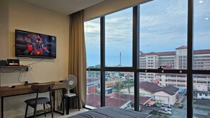 Une chambre d’hôtel avec une grande fenêtre, un lit, un bureau avec une chaise, une télévision et un ventilateur.
