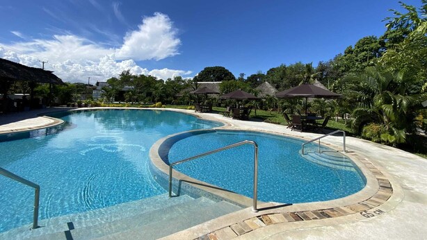 A large, clear swimming pool with a curved design, surrounded by lounge chairs and umbrellas, set in a tropical environment with lush greenery and a clear sky. - White Peacock Resort (Mtwapa)
