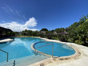 A large, clear swimming pool with a curved design, surrounded by lounge chairs and umbrellas, set in a tropical environment with lush greenery and a clear sky. - White Peacock Resort (Mtwapa)