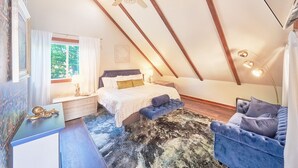 Une chambre avec un plafond en pente, un lit avec une tête de lit, une table de chevet, une chaise et une fenêtre avec des rideaux.