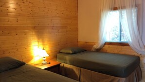 Une chambre aux murs en bois, avec deux lits simples dotés de draps verts, une table de chevet avec une lampe et une fenêtre ornée de rideaux légers.