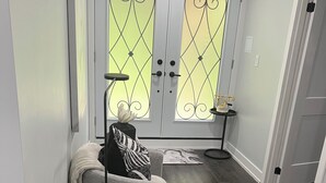 Ein Flur mit einem grau gepolsterten Sessel, einem kleinen Tisch mit einer Lampe und einer dekorativen Tür mit Glasfeldern.