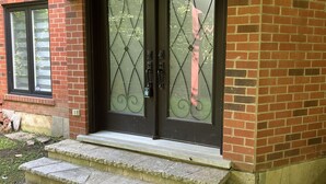 Ein Backsteinhaus mit einer schwarzen Tür, verziert mit dekorativen Glasscheiben und einem Metallgitter. Steinstufen führen zur Tür.