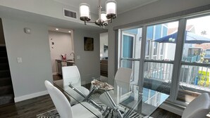 A modern dining area with a glass table and white chairs, a chandelier, and a large window with a view of the outside. - 7 Minute Walk to Beach- Completely Renovated and Pool Views! Two Heated Pool! (Madeira Beach)