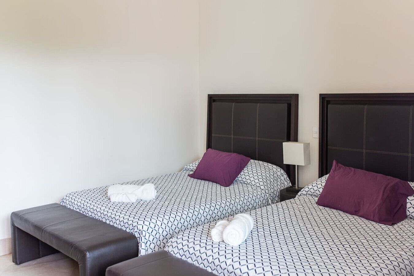 Ein Hotelzimmer mit zwei Betten, jedes mit einem gemusterten Überwurf und einem Kissen. Am Fußende der Betten steht eine Bank und ein Nachttisch mit einer Lampe.