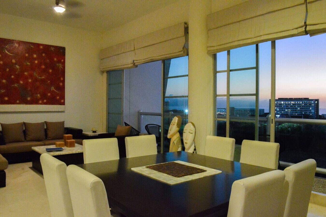 Ein modernes Wohnzimmer mit einem großen Esstisch, weißen Stühlen, einer Couch und Blick auf die Stadt bei Dämmerung.