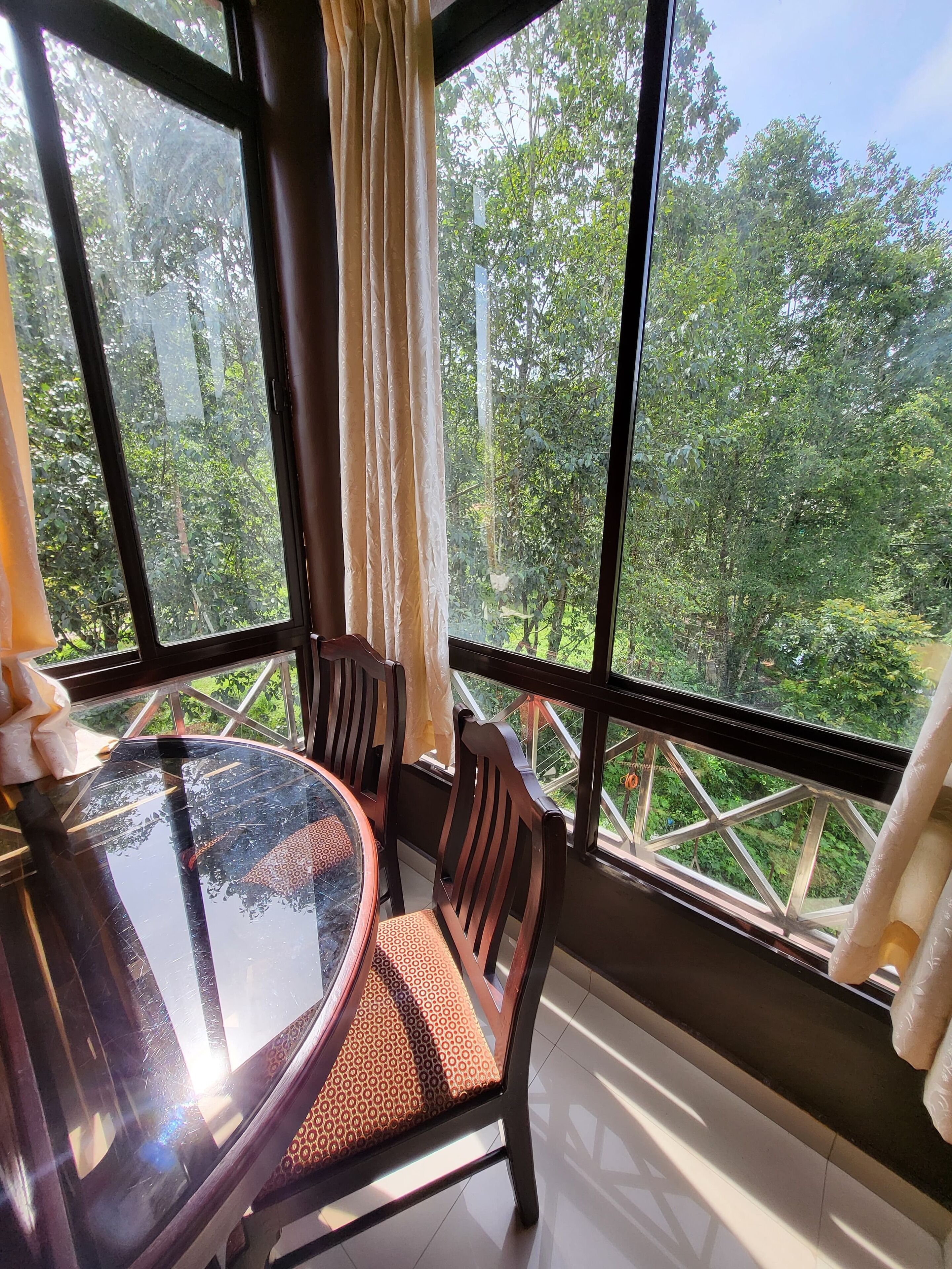 A room with a round glass table and four chairs, large windows with curtains, and a view of greenery outside.