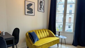 Un sofá amarillo con un cojín azul, una mesita redonda pequeña, una silla y dos cuadros enmarcados en la pared.