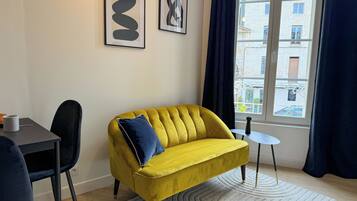 A yellow sofa with a blue pillow, a small round table, a chair, and two framed artworks on the wall.