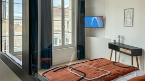 Un dormitorio moderno con un ventanal grande, una cama con colcha estampada, una mesita de noche con un florero y un televisor montado en la pared.