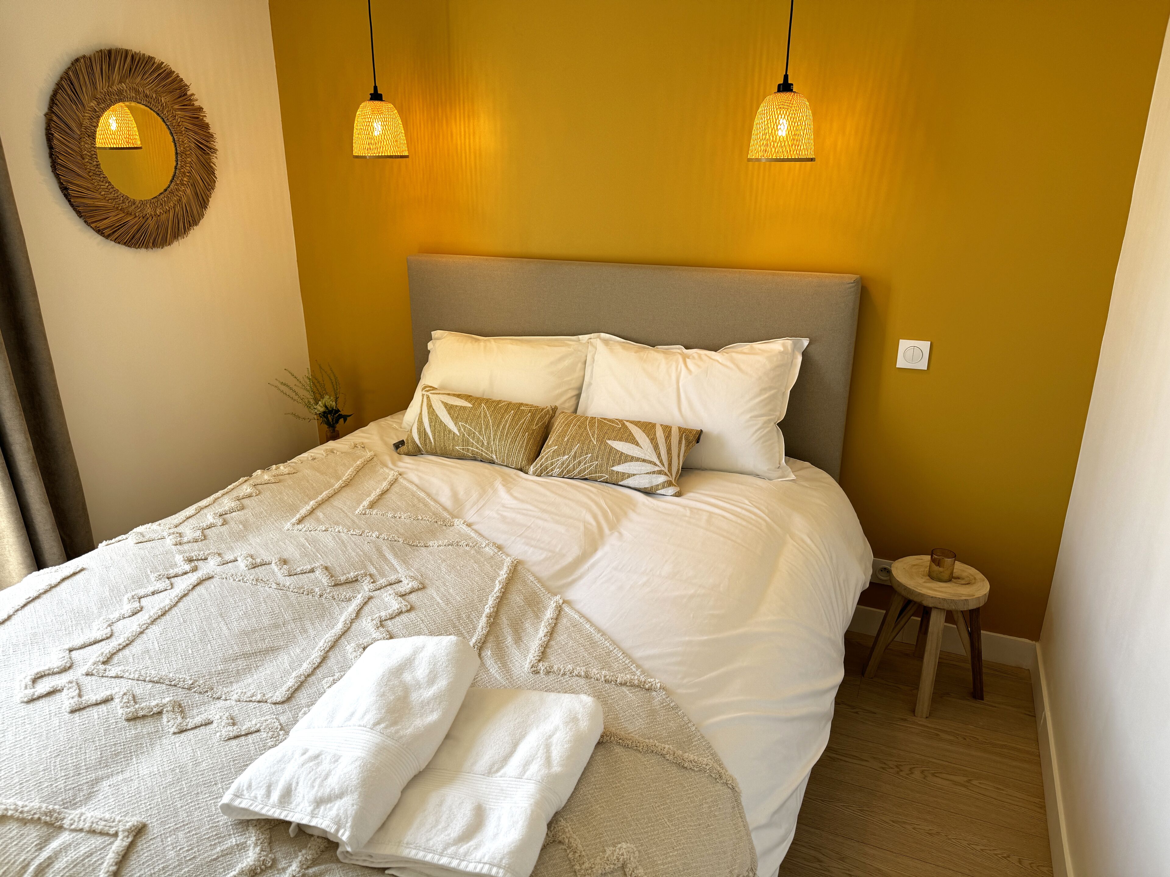 Un lit bien fait, avec du linge de lit blanc et des coussins décoratifs, une table de chevet avec un verre, et un miroir fixé au mur.
