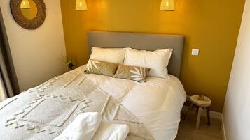 A neatly made bed with white linens and decorative pillows, a bedside table with a glass, and a wall-mounted mirror.