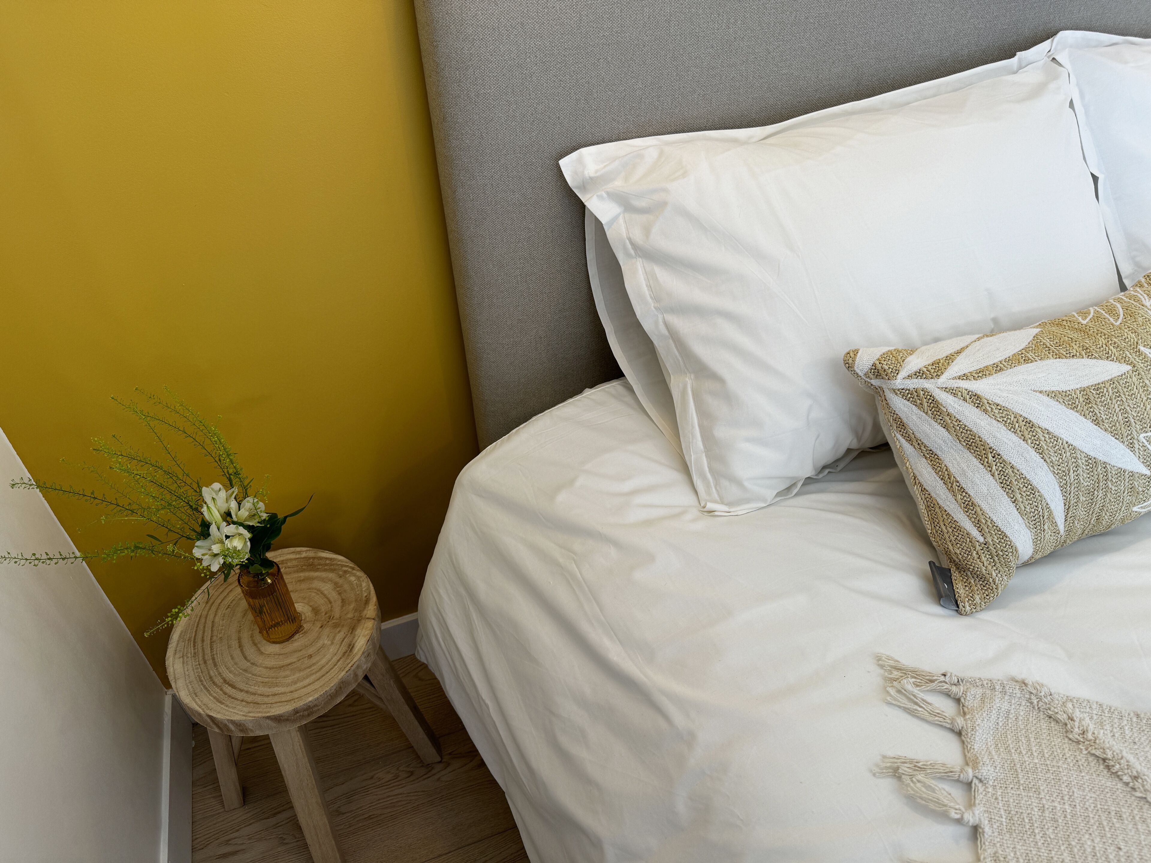 A neatly made bed with white bedding, a grey headboard, and a small round wooden stool with a woven top and a vase of flowers beside it.