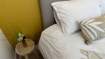 Un letto rifatto con lenzuola bianche, una testiera grigia e uno sgabello rotondo in legno con un piano intrecciato, con un vaso di fiori accanto.