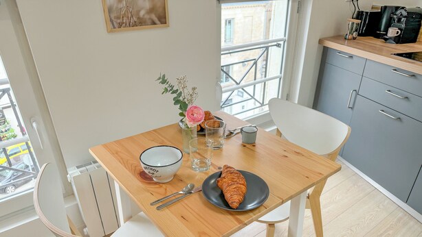 Ein Essbereich mit einem Holztisch, weißen Stühlen, einem Teller mit einem Croissant, einer Schüssel und einer Vase mit Blumen.