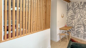 Ein Raum mit einer Trennwand aus Holzleisten, einem kleinen Holztisch mit Stuhl, einer Wand mit Blattmuster und einem grünen Sofa.