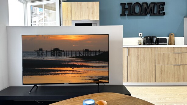 Een moderne keuken met een flatscreen-tv waarop een zonsondergang te zien is, houten keukenkastjes en een bordje met de tekst 'HOME'.