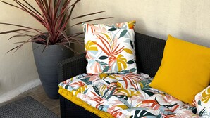Een rieten bank met een kleurrijk bloemenkussen en een geel kussen, een hoge potplant met lange bladeren, en een getextureerd vloerkleed op een betegelde vloer.
