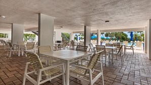 An outdoor patio area with tables and chairs, a pool in the background, and a view of palm trees.