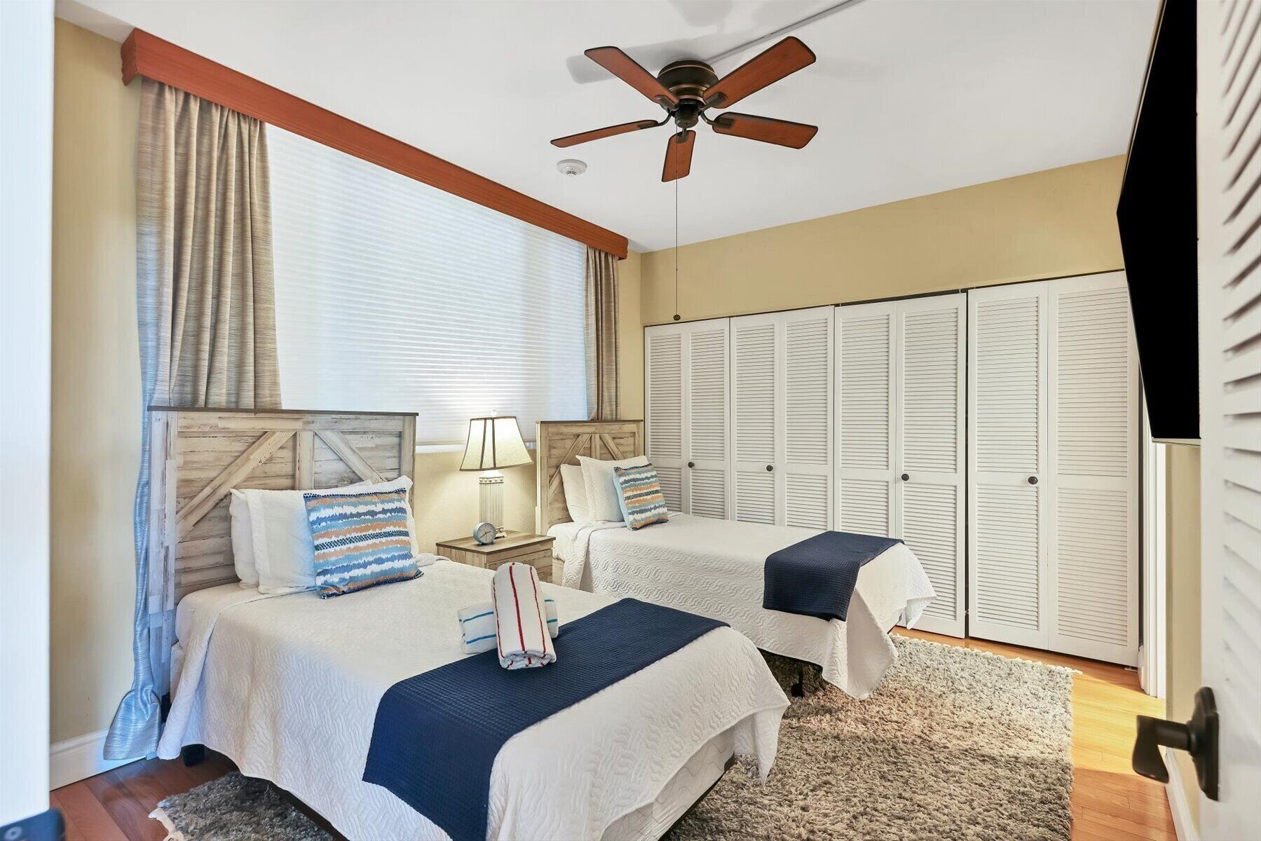 A bedroom with two beds, a ceiling fan, a window with curtains, and a set of white closets.